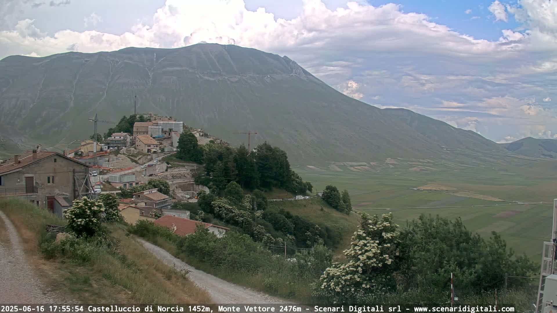 A partly cloudy day reveals a hillside village nestled below a large mountain, with construction visible in the village.
