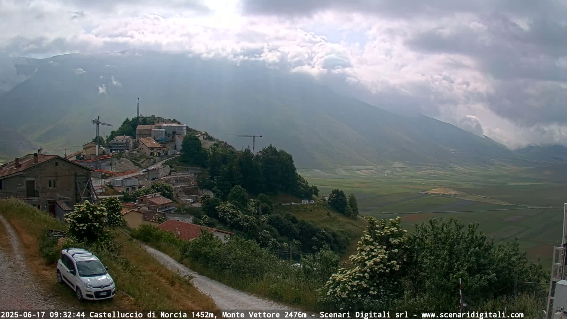 A hillside village with construction equipment is visible beneath a cloudy sky, overlooking a vast, flat valley.