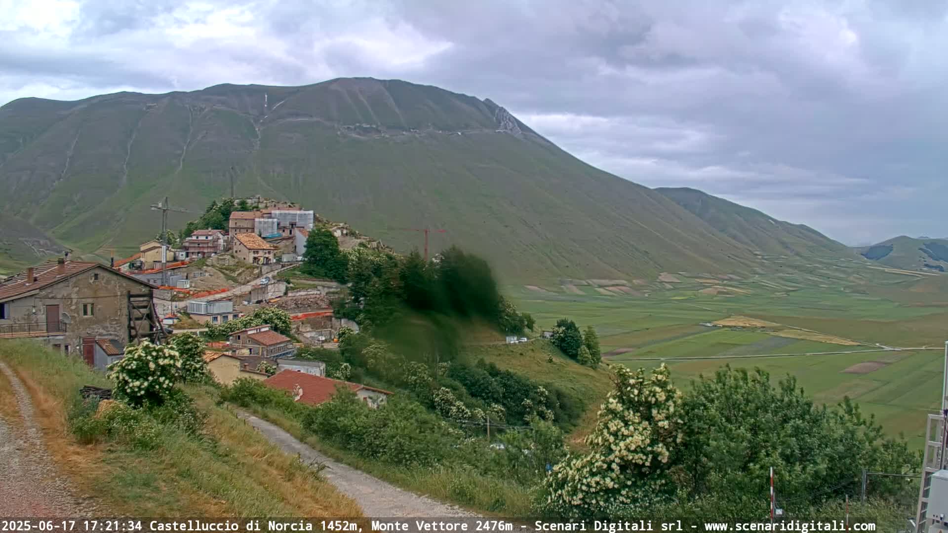 A hilltop village nestled against a large mountain under a cloudy sky overlooks a vast, verdant plain.