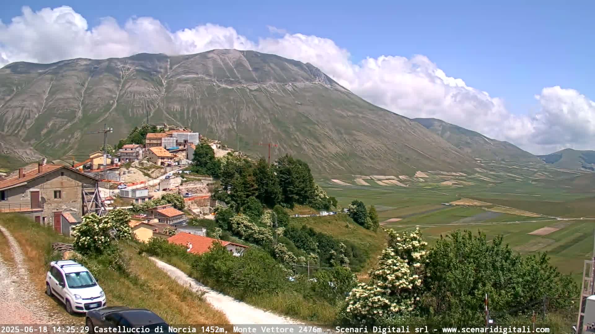 A mountain village nestled against a large, grey mountain under a partly cloudy sky, with a valley of farmland stretching out below.