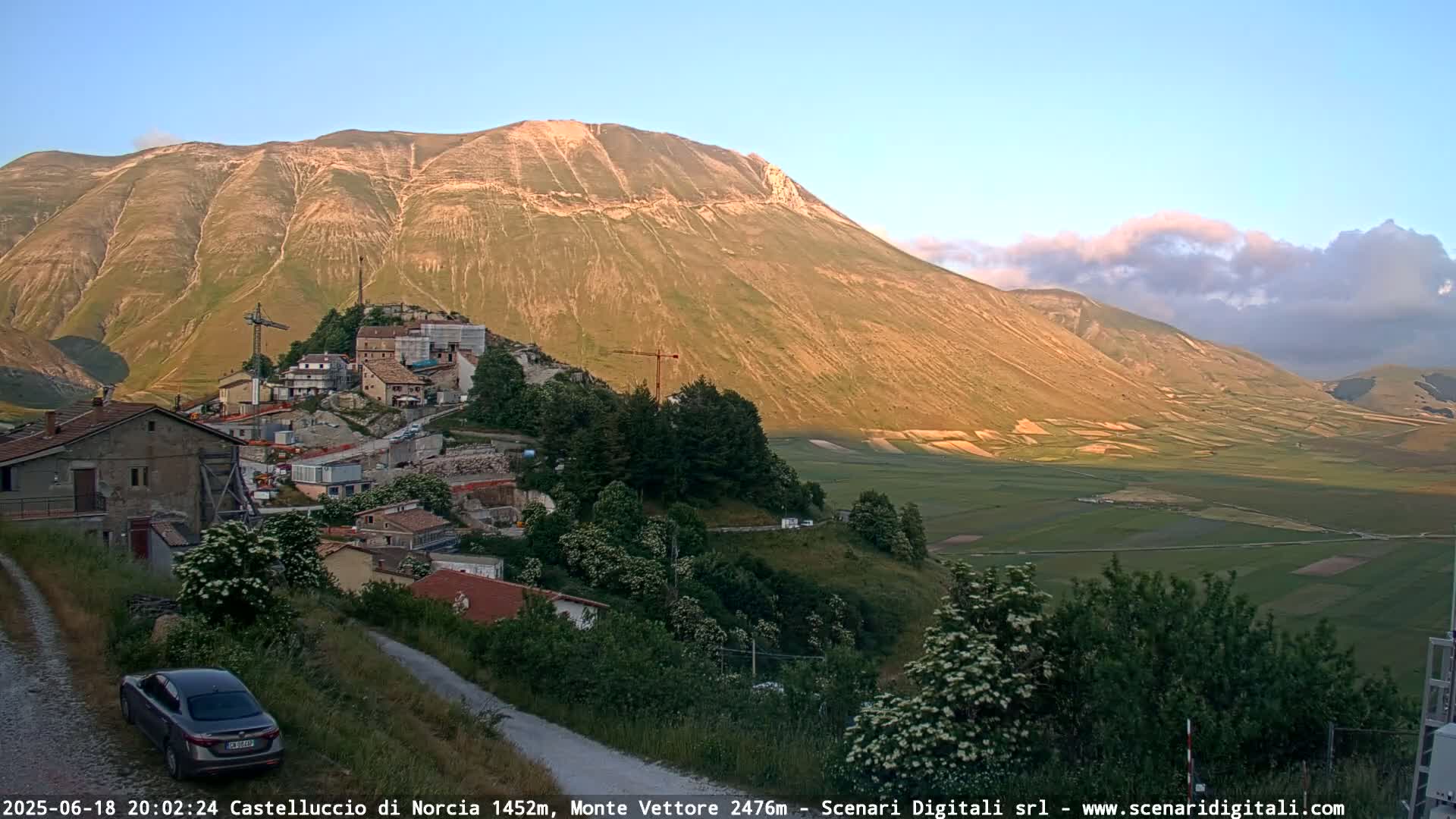 A mountain village nestled in a valley below a large, sunlit mountain under a partly cloudy evening sky.