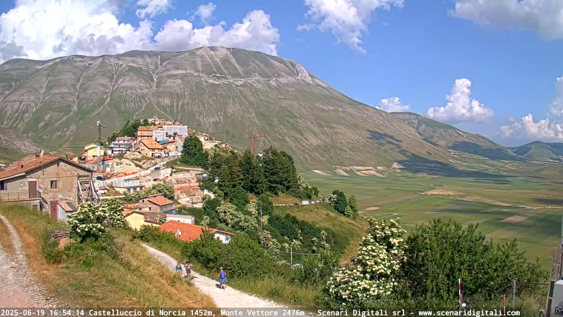 A partly sunny day reveals a mountain village nestled below a large mountain range, with a valley of farmland stretching out in the distance.