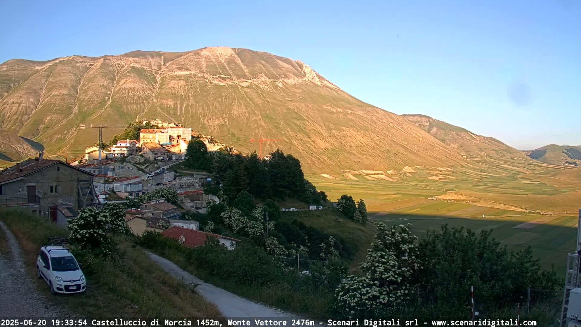 A mountain village nestled against a large, sunlit mountain overlooks a valley of farmland under a clear sky.