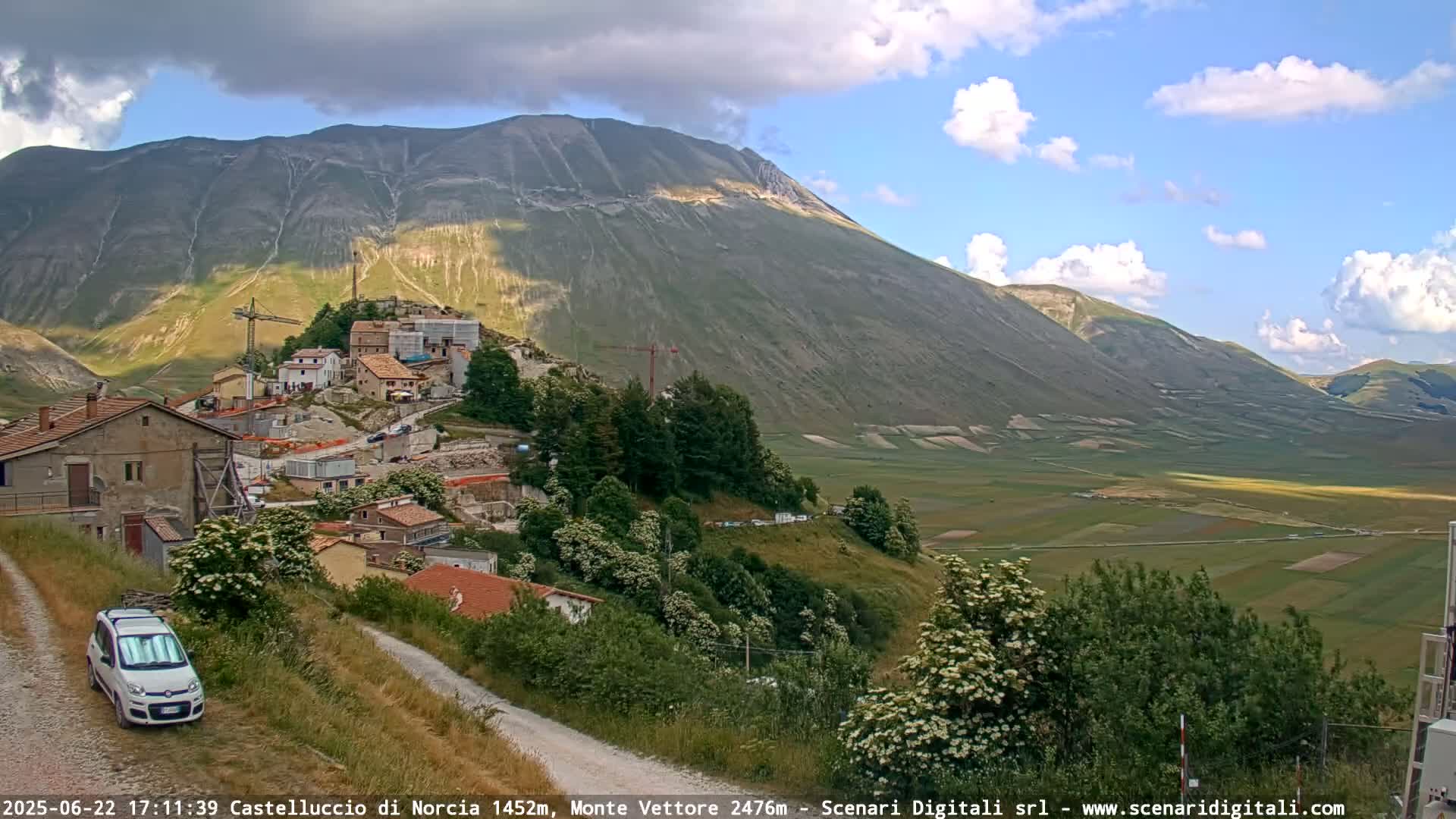 A partly cloudy day reveals a village nestled on a hillside below a large mountain, with a valley of farmland stretching out in the distance.