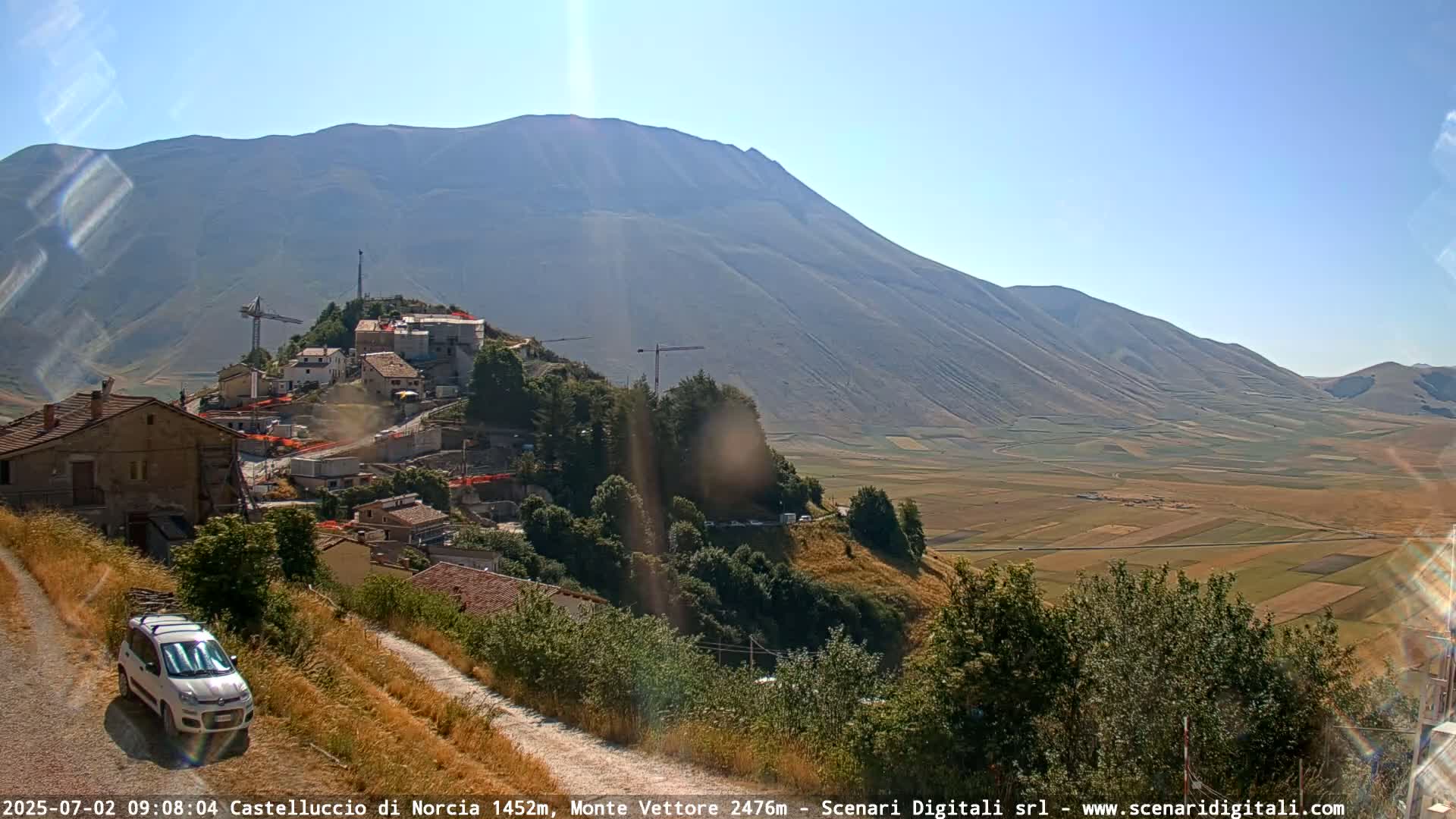 A sunny, clear day reveals a hillside town undergoing construction, nestled below a large mountain and overlooking a vast, flat agricultural valley.