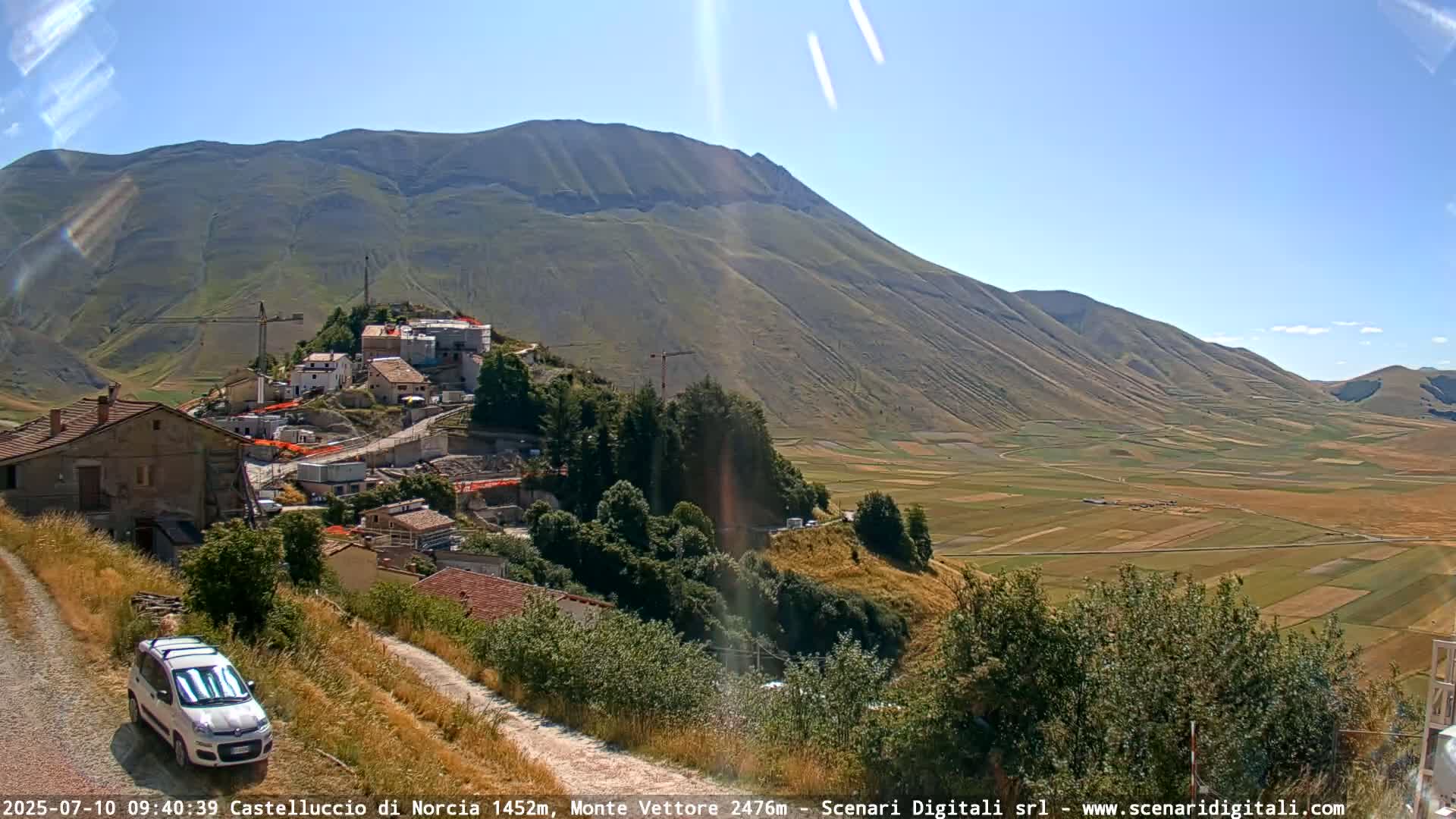 A small village nestled on a hillside overlooks a vast, flat valley of farmland under a clear, sunny sky, with construction visible in the village.