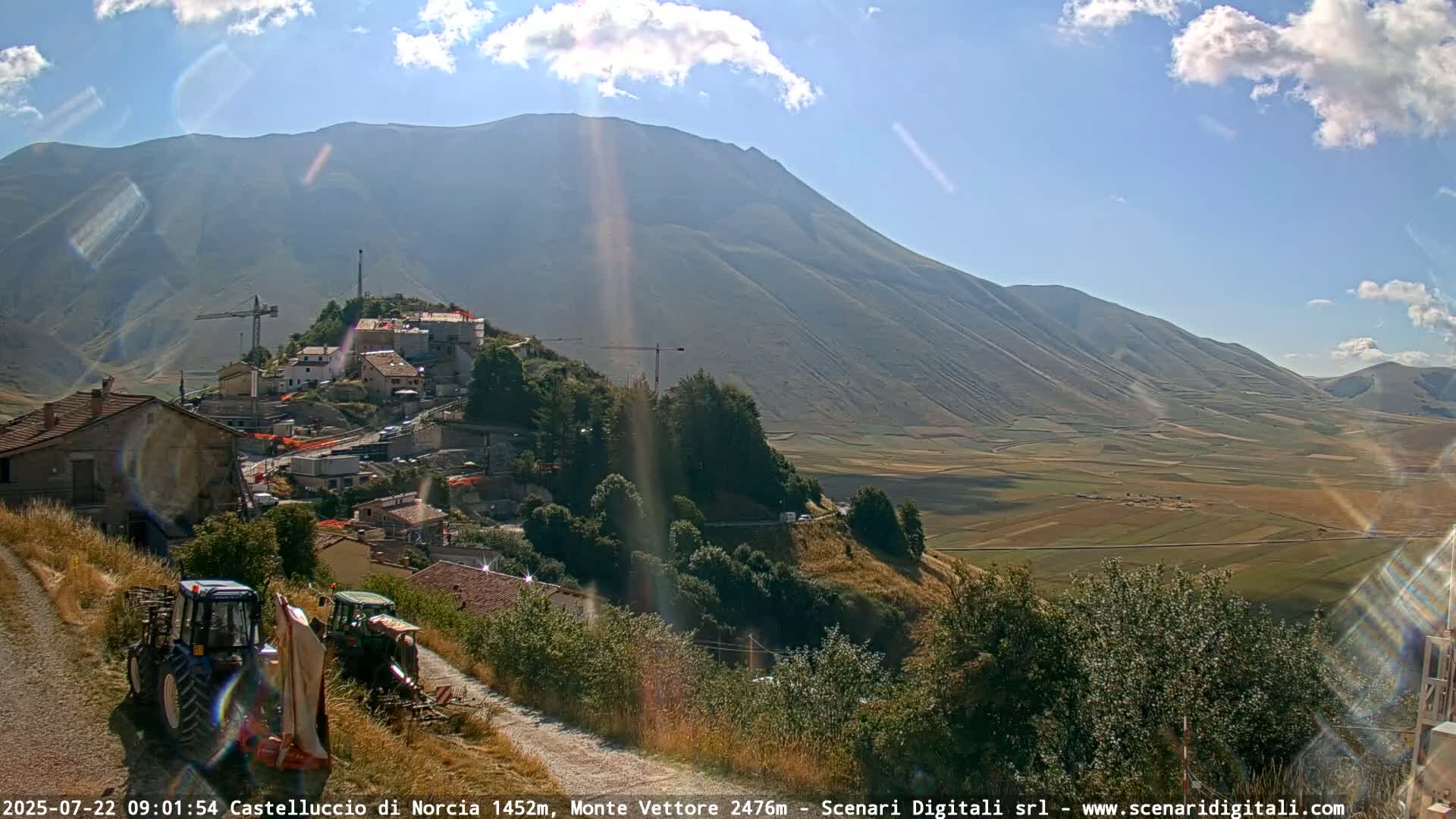 A sunny day reveals a hillside village nestled beneath a large mountain, with a valley of farmland stretching into the distance, and two tractors parked on a nearby road.