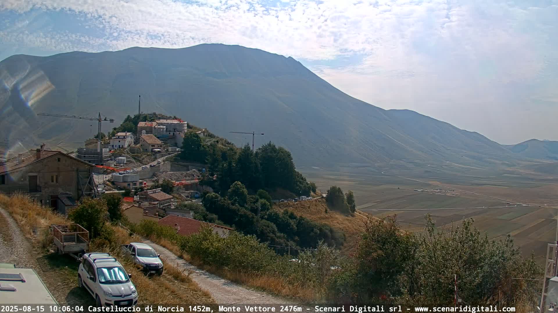 A partly cloudy day reveals a mountain valley with a small town under construction, viewed from a slightly elevated vantage point showing two cars parked on a dirt road.