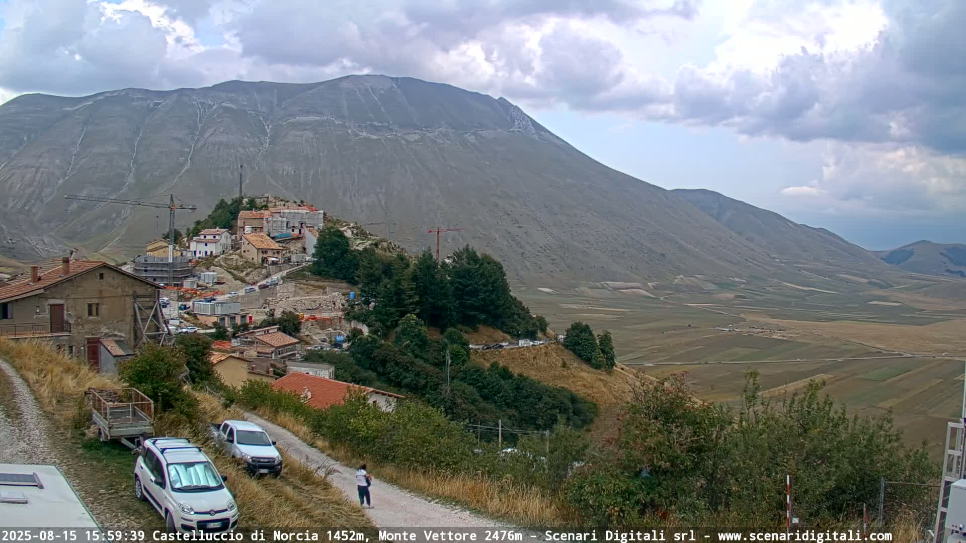 A partly cloudy day reveals a mountain valley with a small town under construction, viewed from a slightly elevated vantage point showing two cars parked on a dirt road.
