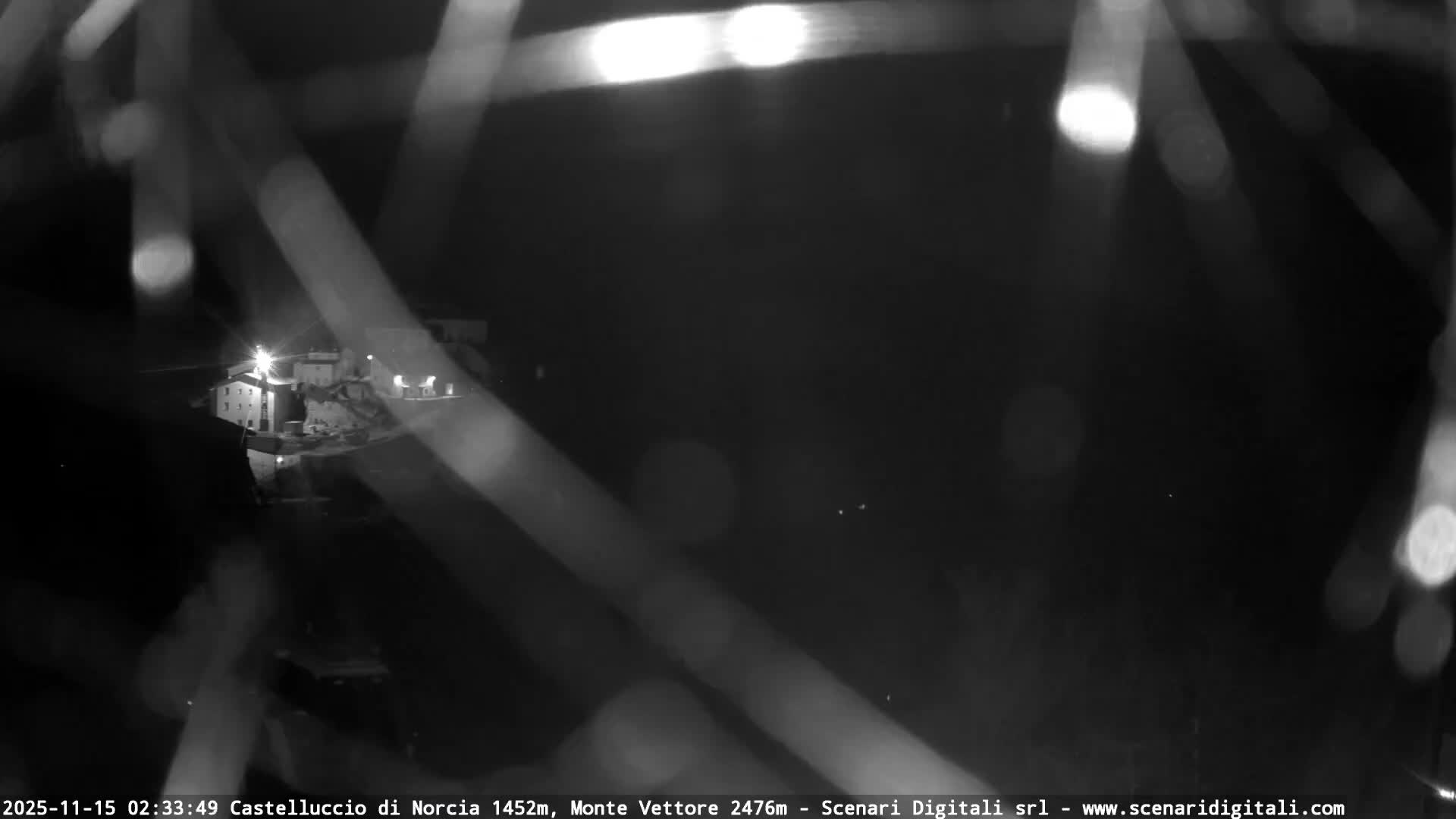 A blurry, monochrome, nighttime webcam view shows a brightly lit village on a distant hillside, partially obscured by heavy precipitation (likely rain or snow) close to the lens, indicating inclement weather.