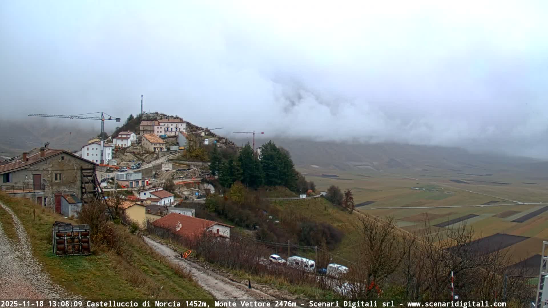 A blurry, monochrome, nighttime webcam view shows a brightly lit village on a distant hillside, partially obscured by heavy precipitation (likely rain or snow) close to the lens, indicating inclement weather.