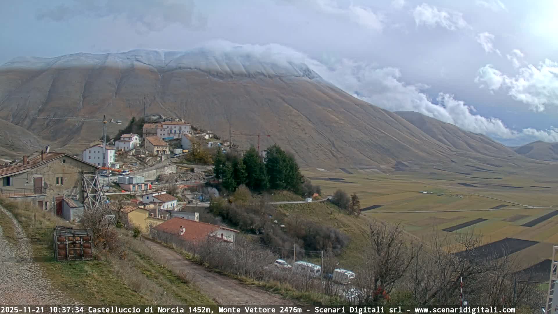 A hillside village with signs of construction overlooks a vast valley of agricultural fields, backed by large, partly cloud-covered mountains under a partly cloudy sky.