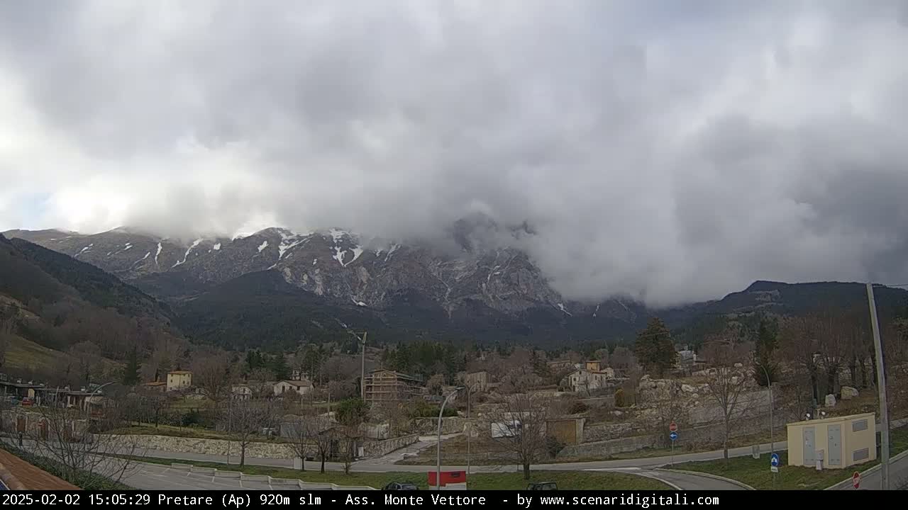 A small town nestled in a valley, with snow-capped mountains shrouded in clouds in the background under an overcast sky.
