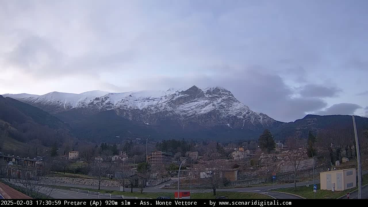 A snow-capped mountain range rises behind a small town under a mostly cloudy sky.