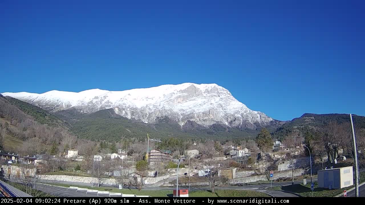 A snow-capped mountain rises behind a small village nestled in a valley under a clear blue sky.