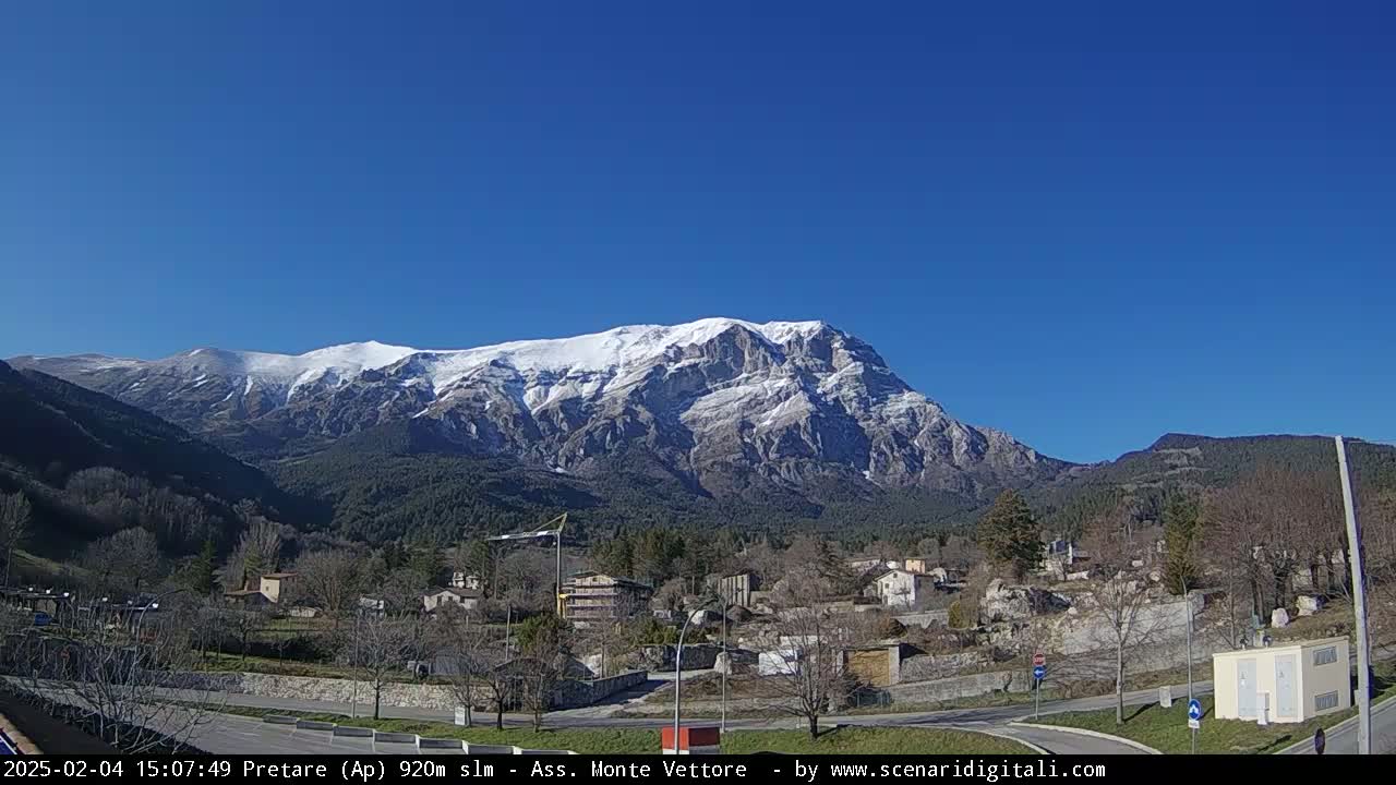 A snow-capped mountain range looms over a small town nestled in a valley on a clear, sunny day.