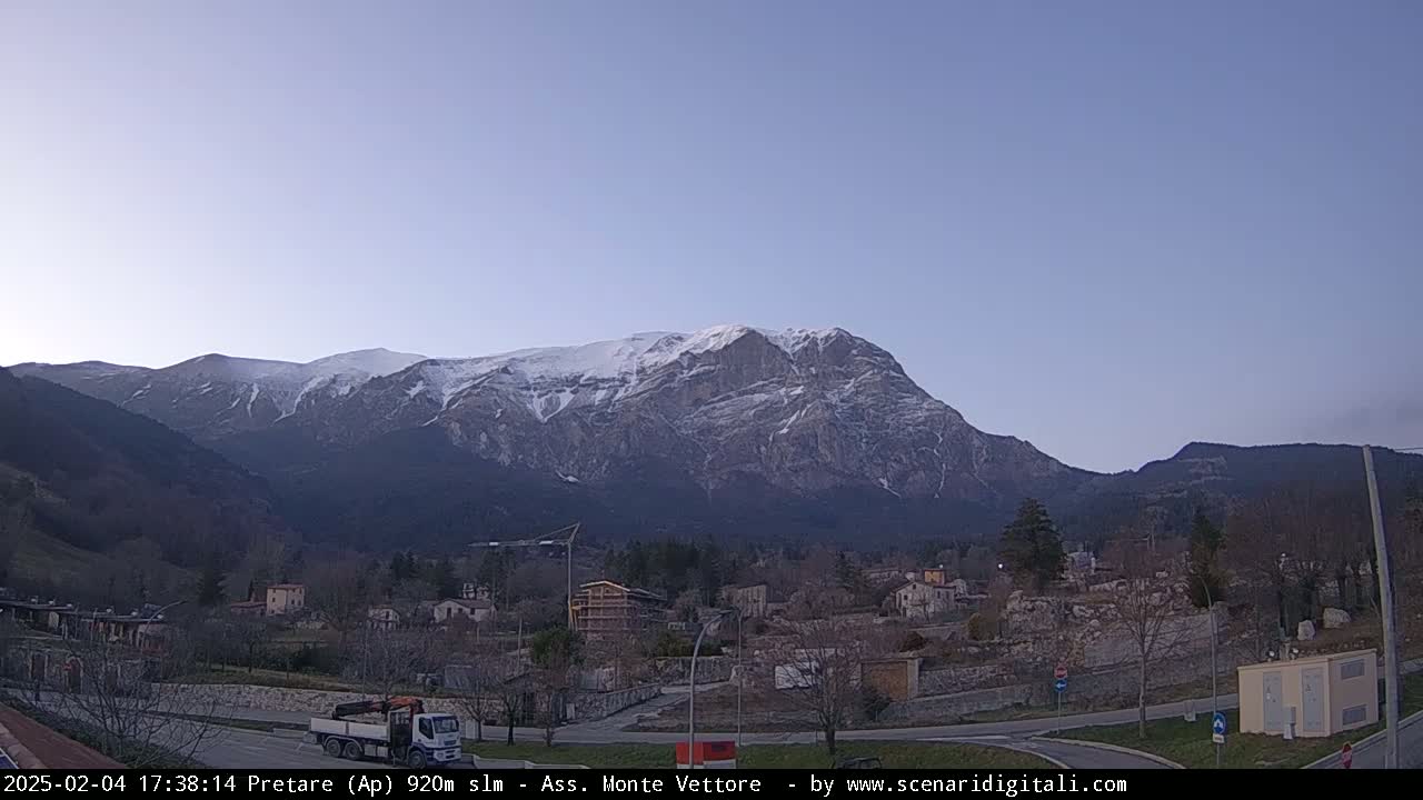 A snow-capped mountain looms over a small town nestled in a valley under a clear, twilight sky.