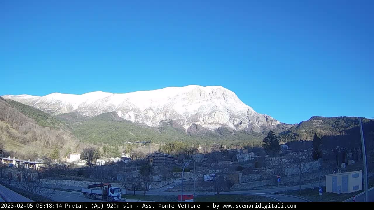 A snow-capped mountain rises above a small town nestled in a valley on a clear, sunny day.