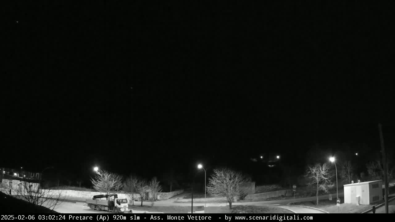 A nighttime, clear-sky view of a mostly empty roadway with a few bare trees, a truck, and a small building.
