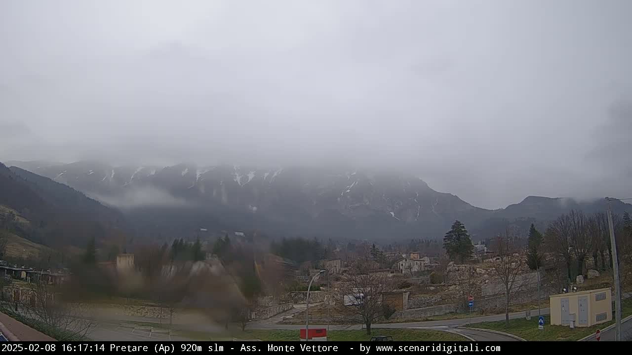 A foggy, overcast day reveals snow-dusted mountains in the distance, overlooking a small town with sparse trees and buildings.