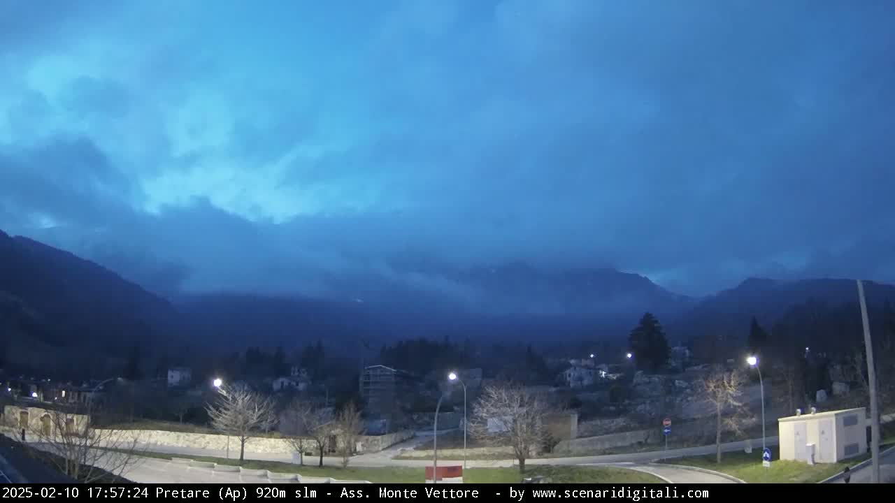 A nighttime view of a valley town nestled between mountains under a cloudy, dark blue sky.