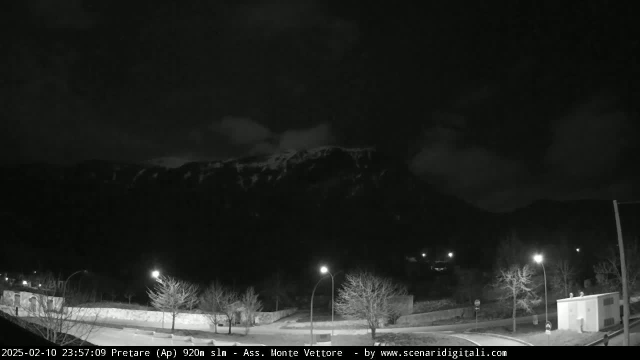 A nighttime, cloudy scene shows a snow-capped mountain range in the distance, with dimly lit roads and trees in the foreground.