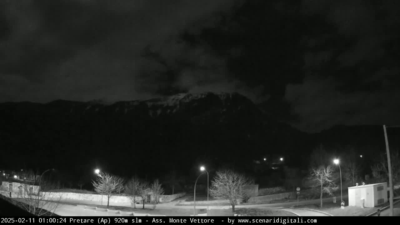 A nighttime view of a snow-capped mountain range under a dark, cloudy sky, with sparsely lit roads and trees in the foreground.