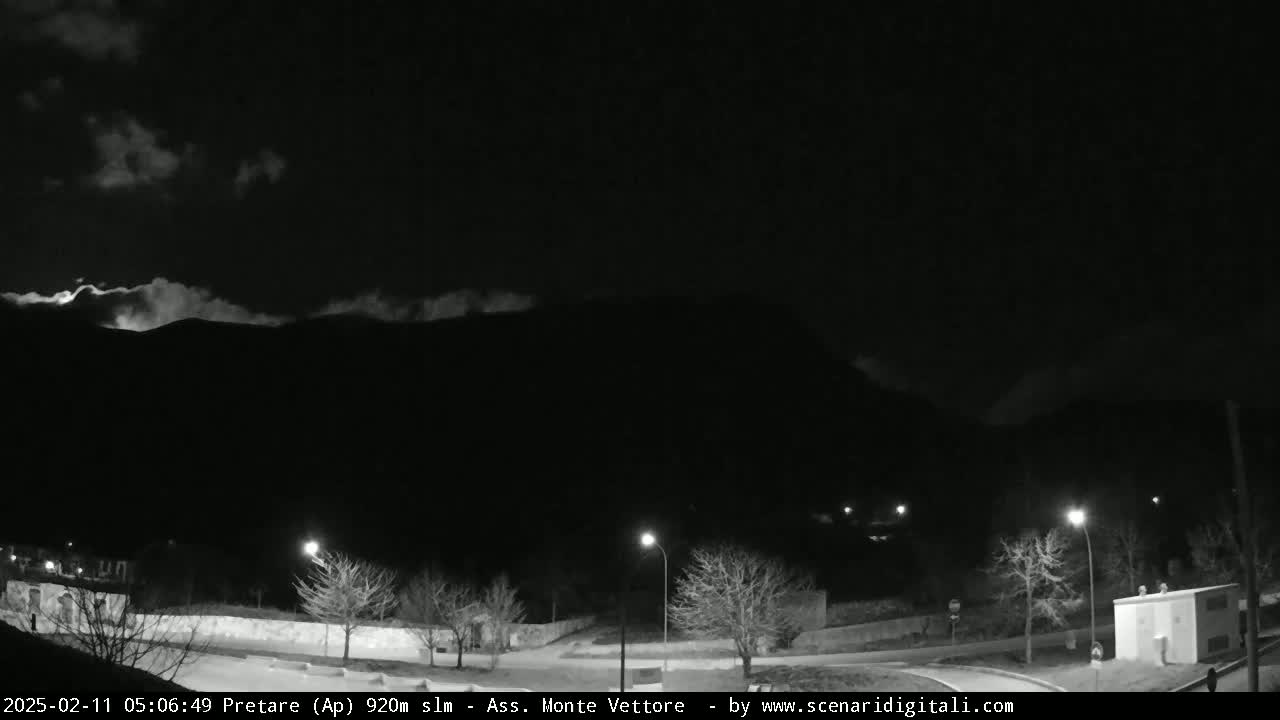 A nighttime, black-and-white view of a mountain range under a partly cloudy sky, with a small town and streetlights visible in the foreground.
