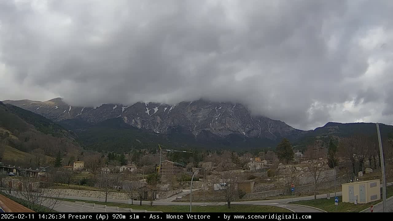 A small town nestled in a valley is overshadowed by a large, snow-dusted mountain range under a cloudy sky.