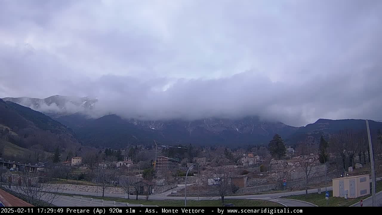 A town nestled in a valley is partially obscured by fog and low-hanging clouds, with snow-dusted mountains in the background under an overcast sky.