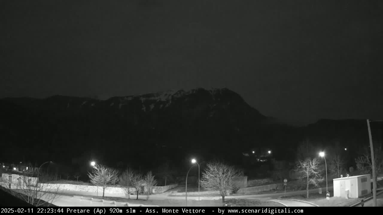 A nighttime view of a snow-dusted mountain looming over a sparsely lit road with leafless trees and a small building.