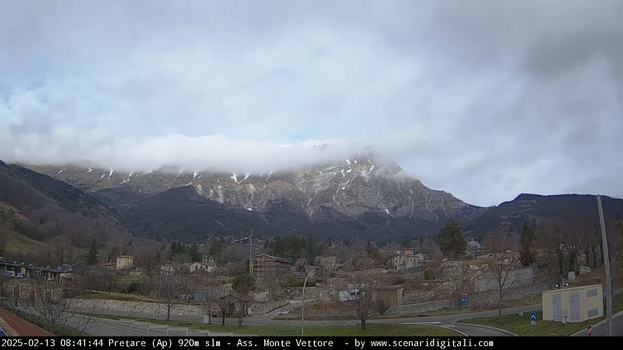 A snow-dusted mountain range is partially obscured by clouds, overlooking a small town in a valley under an overcast sky.
