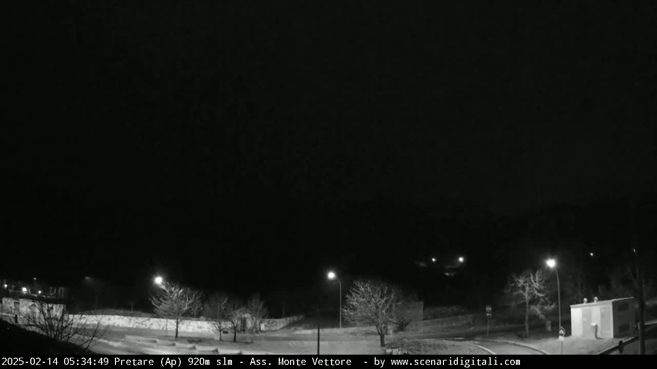 A nighttime, black-and-white view of a sparsely lit road curving through a snow-covered landscape with leafless trees and a small building.
