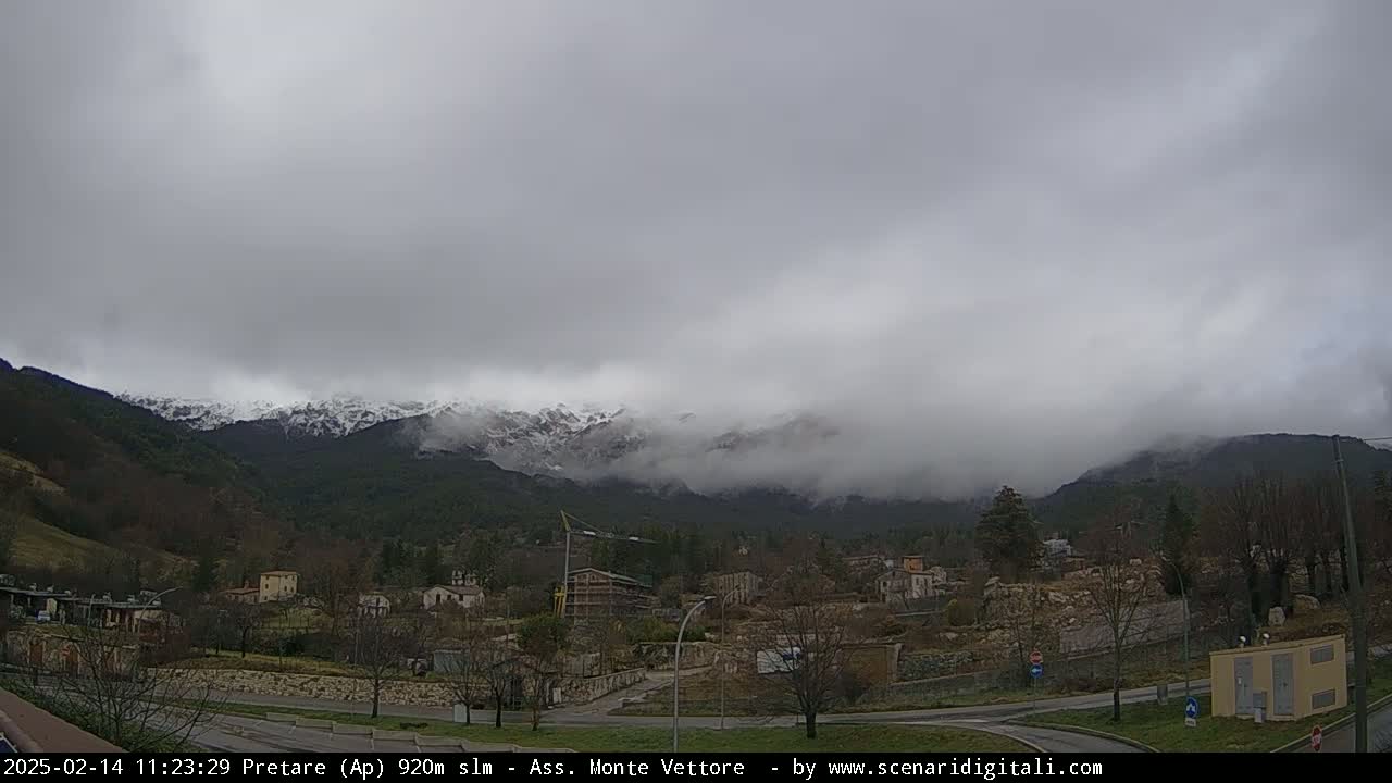 A small town sits in a valley beneath snow-capped mountains shrouded in fog and low-hanging clouds.