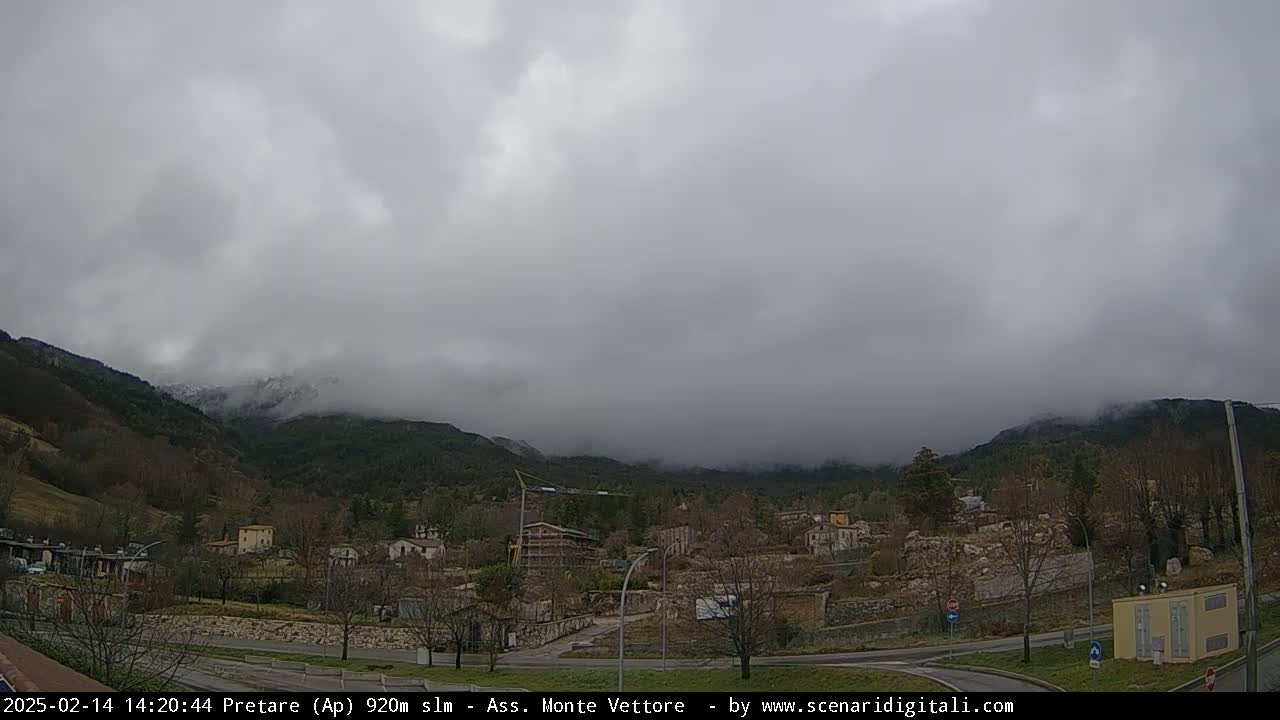 A small town nestled in a valley between overcast mountains, some of which are lightly dusted with snow.