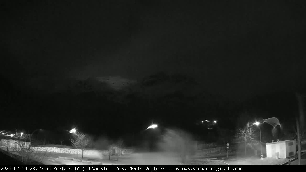 A nighttime, snowy scene shows a road with streetlights illuminating a small building and surrounding area, with blowing snow obscuring some distant features.