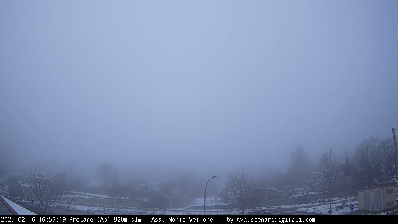A thick fog obscures a sparsely populated landscape with bare trees and a few buildings, some patches of snow are visible on the ground.