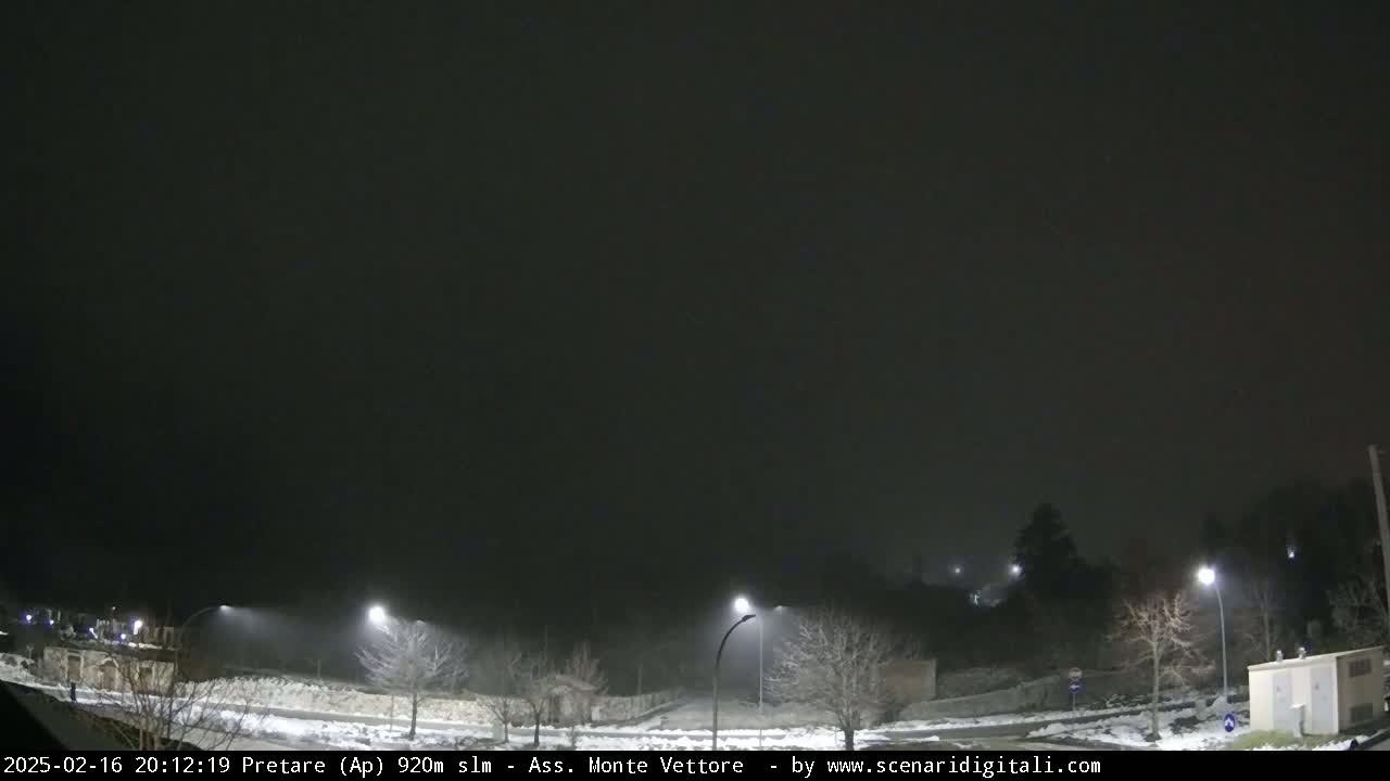 A snow-covered road at night is dimly lit by streetlights in foggy conditions.