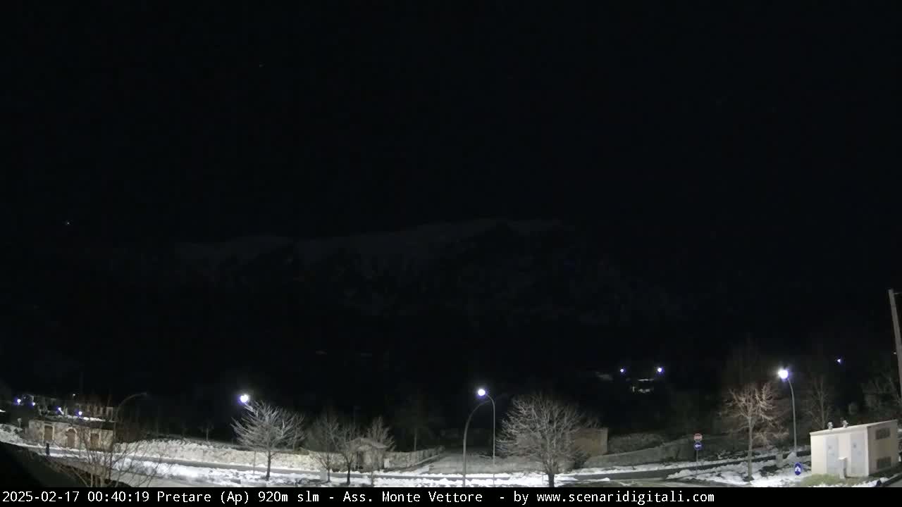 A nighttime view of a snow-covered road and sparsely populated area with streetlights, under a dark, clear sky, shows a mountain range in the distance.