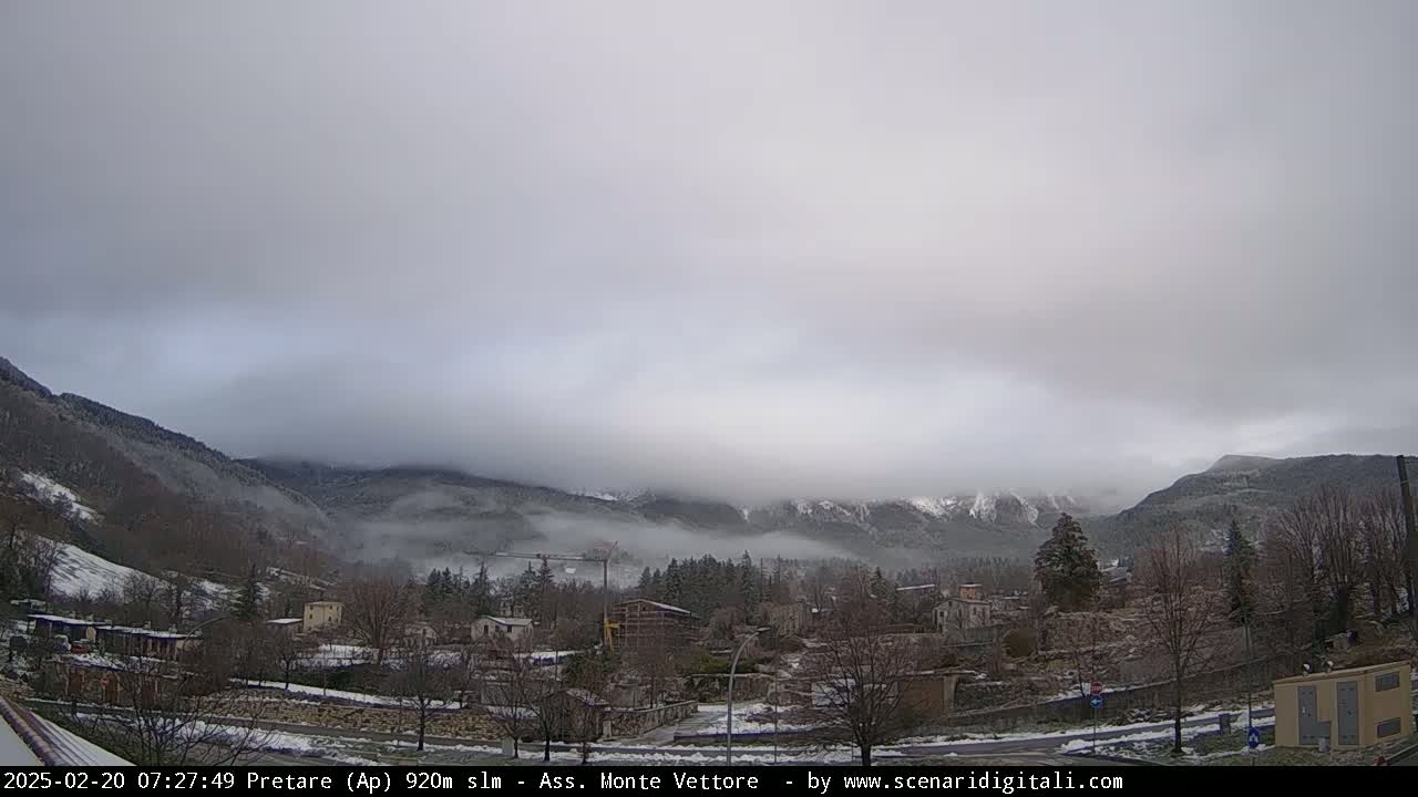 A valley town sits beneath a low-hanging overcast sky, with snow dusting the ground and nearby mountains.