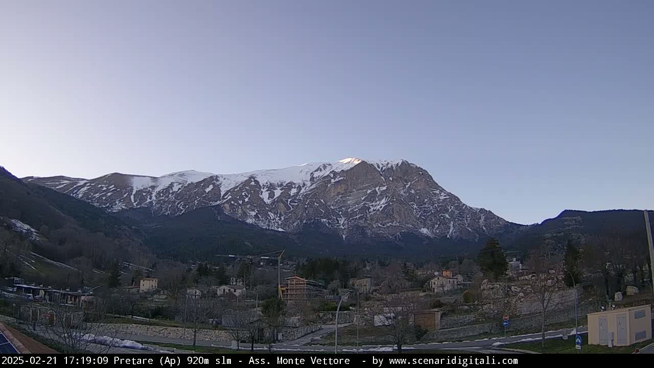 A snow-capped mountain looms over a small town nestled in a valley under a clear, twilight sky.