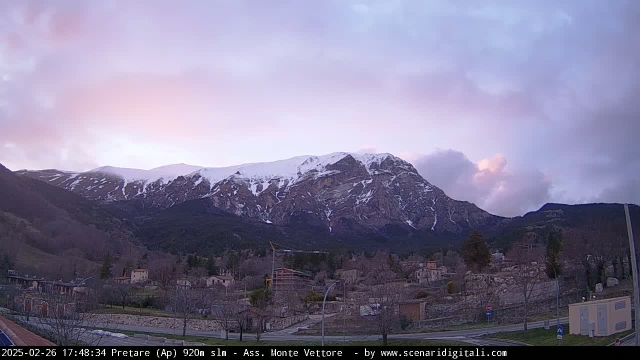 A snow-capped mountain range is visible under a partly cloudy, pinkish-purple twilight sky above a small village.