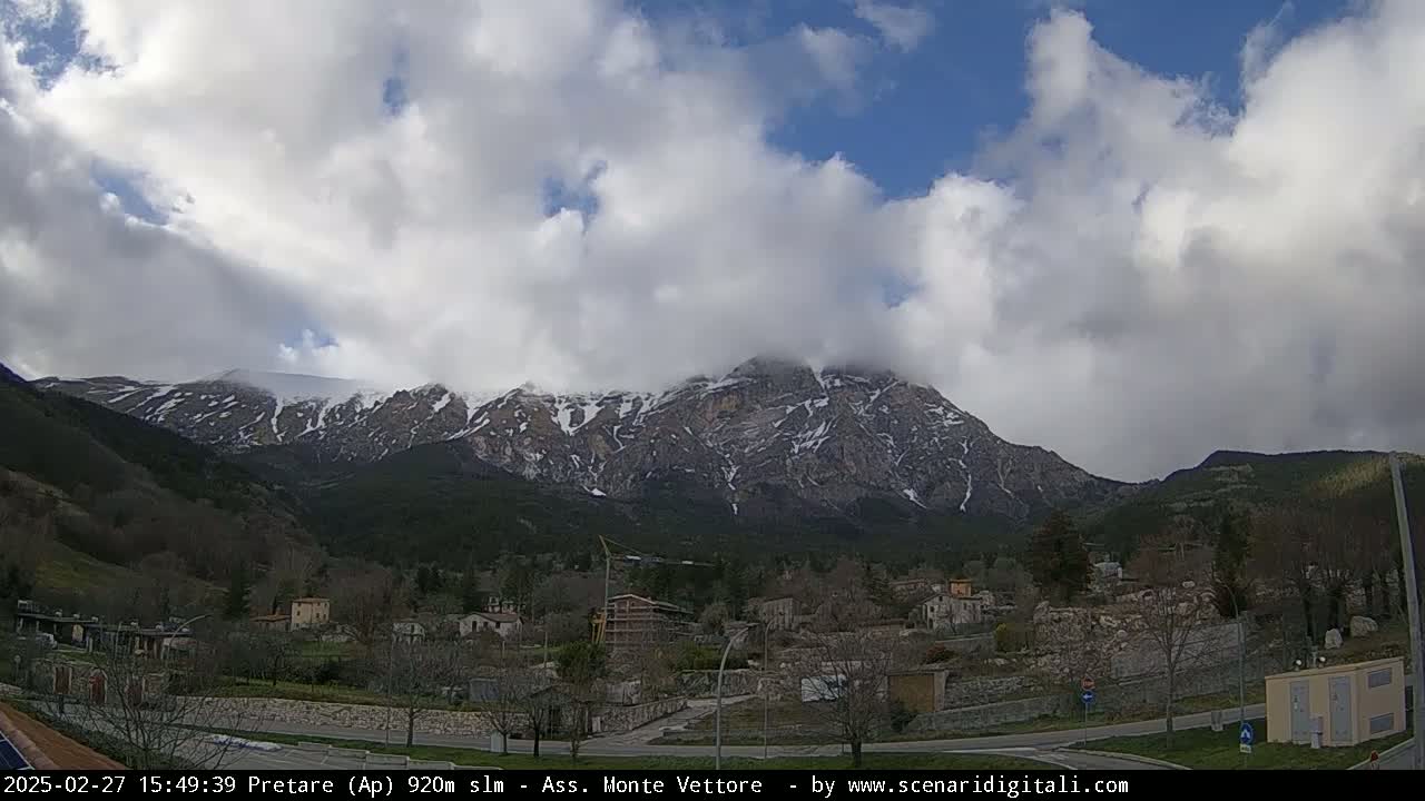 A partly cloudy day reveals a snow-dusted mountain range overlooking a small village nestled in a valley.