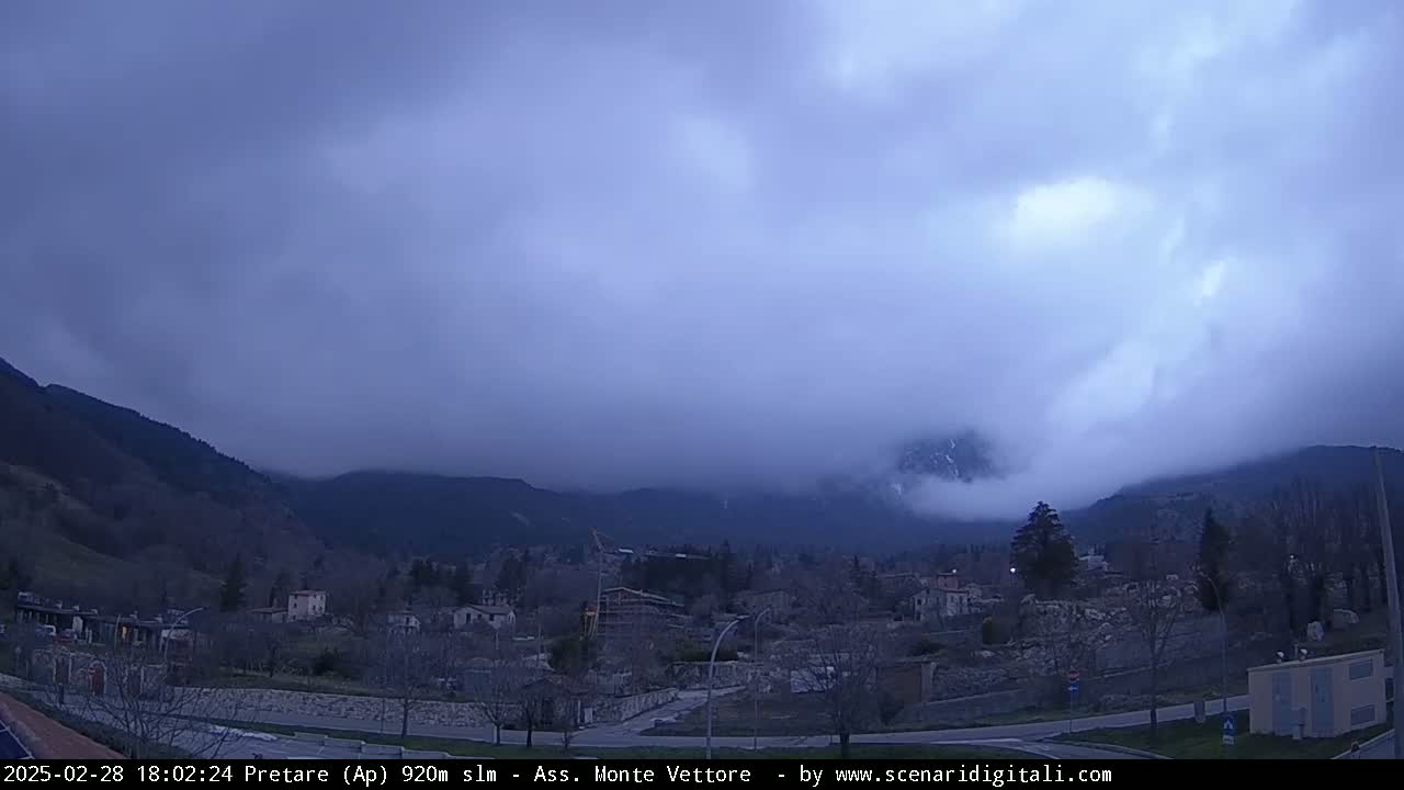 A small town nestled in a valley is shrouded in a thick layer of fog and low-hanging clouds, with mountains rising in the background under a mostly cloudy, dusky sky.