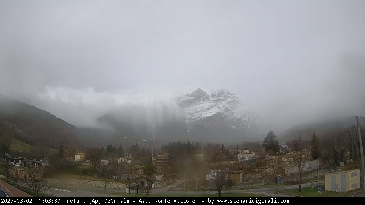 A snow-dusted mountain is partially obscured by heavy fog, overlooking a small village nestled in a valley.