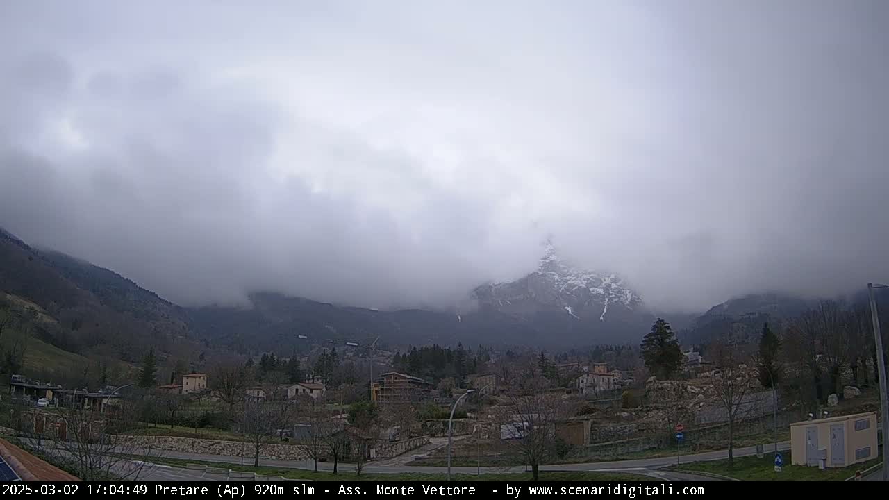 A small town nestled in a valley is shrouded in fog, with a snow-capped mountain partially visible in the background under an overcast sky.