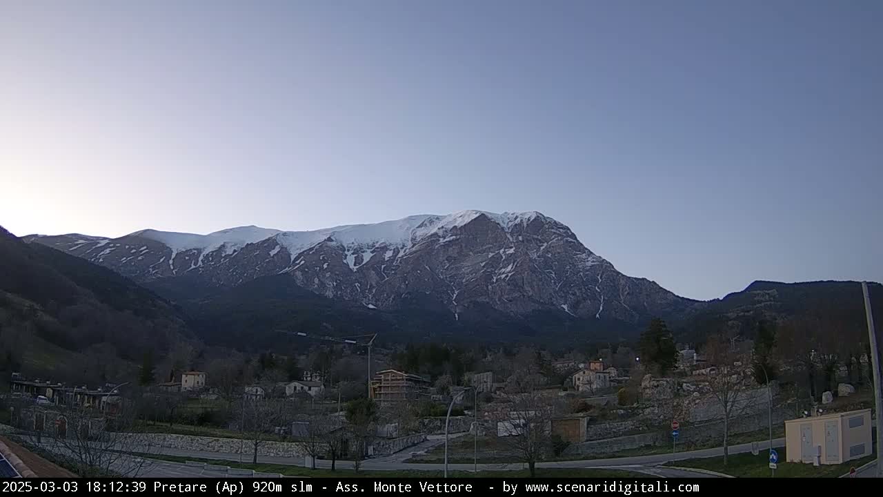 A snow-capped mountain range looms over a small town nestled in a valley under a clear twilight sky.