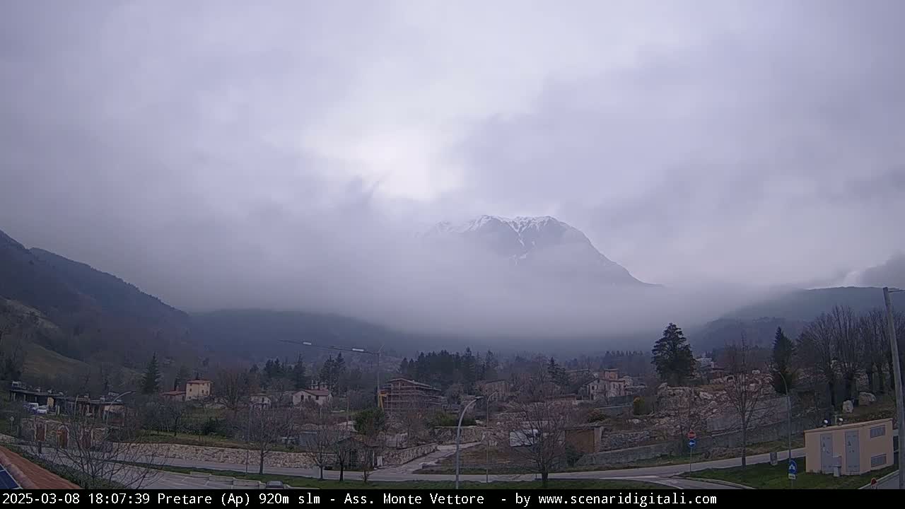 A snow-capped mountain is partially obscured by fog, overlooking a small village nestled in a valley.
