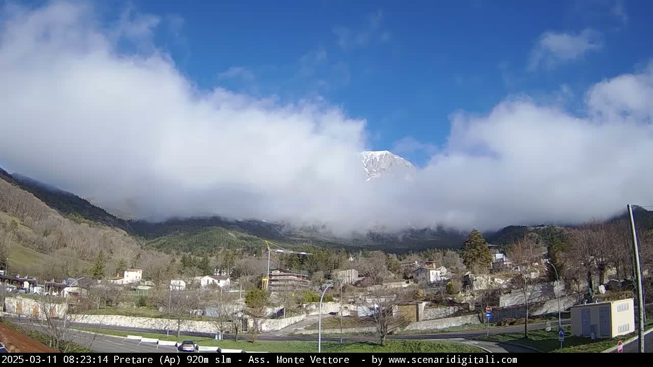 A partly cloudy, sunny day reveals a snow-capped mountain peak partially obscured by clouds, overlooking a small town nestled in a valley.
