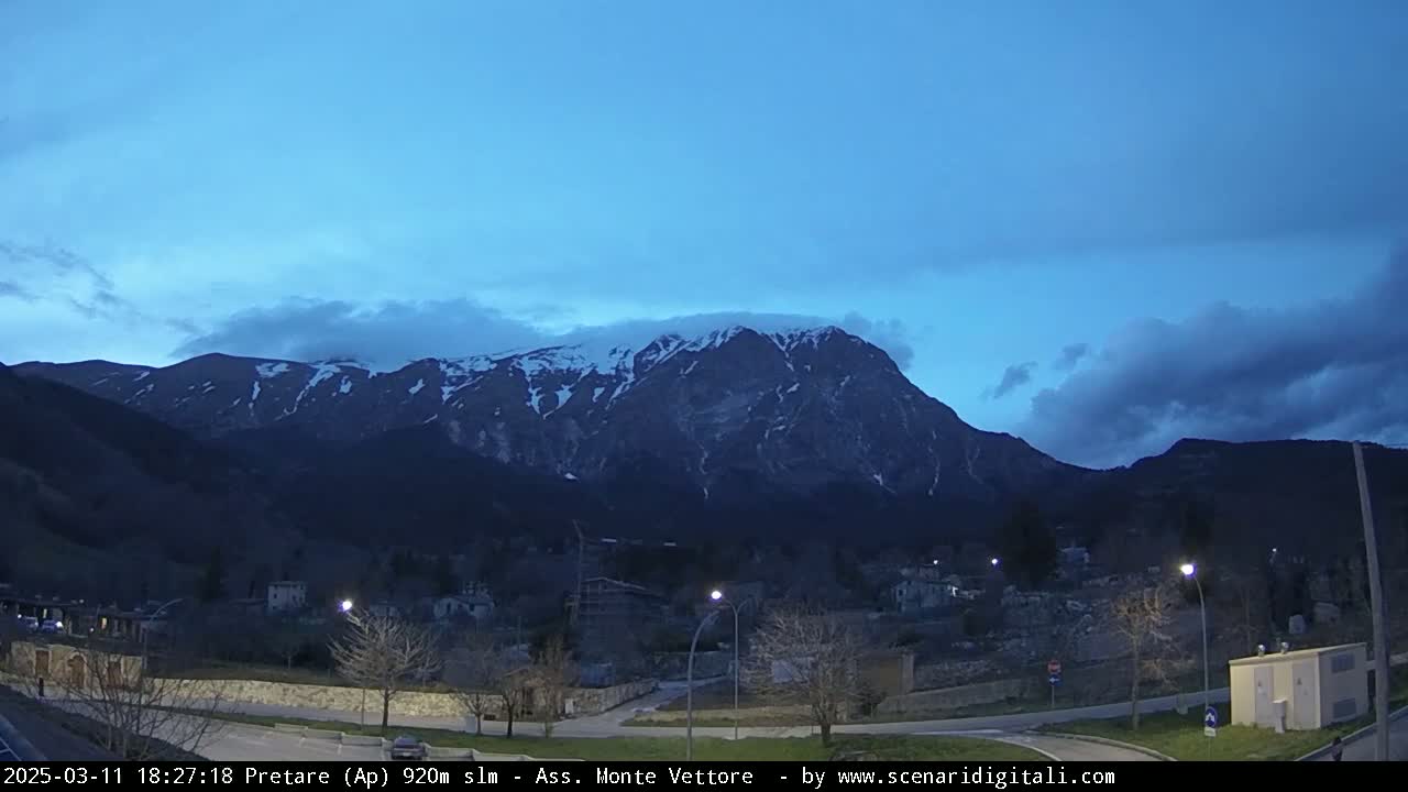 A snow-capped mountain looms over a small town at dusk under a mostly clear, twilight sky.
